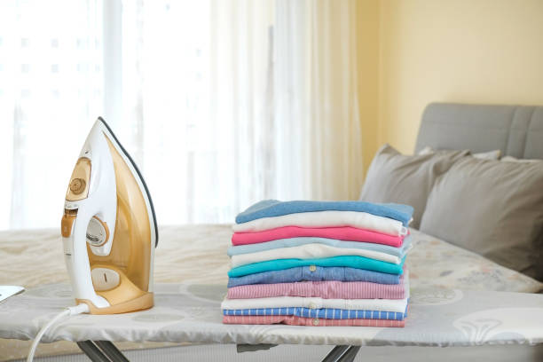 Ironing Board and the Formation of Clothing Aesthetic Standards 2 Stacked folded clothes next to an iron on an ironing board in a bright room.
