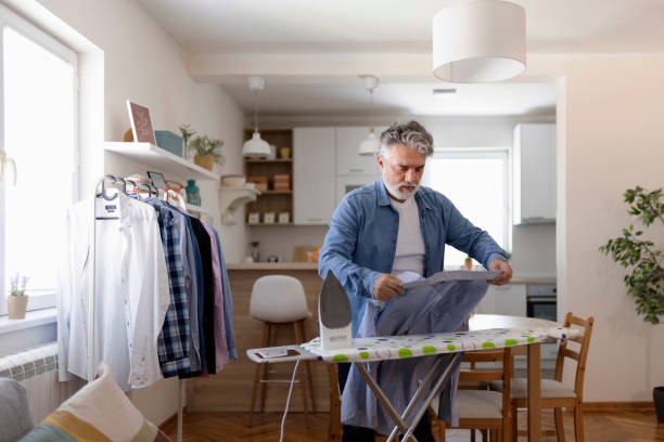 Man with a beard irons a blue shirt on an ironing board in a clean, modern home.