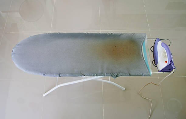 Empty, well-used grey ironing board with a purple iron on its stand.