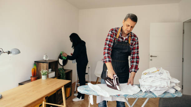 Man in apron irons clothes on a polka-dot ironing board; woman cleans in background.