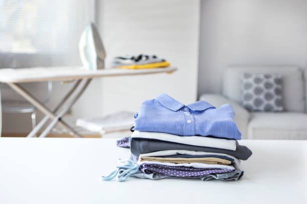 Stack of clean, folded laundry on a table, with an ironing board behind.