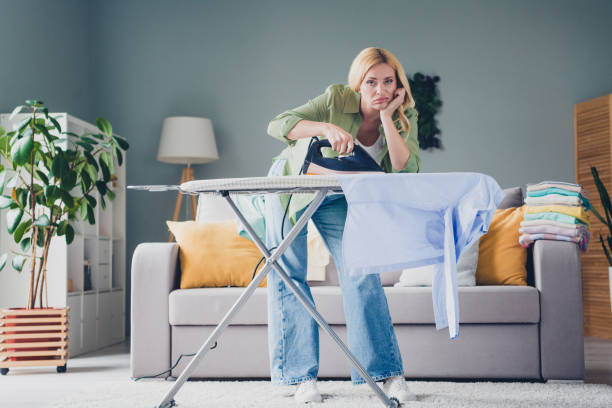 Tired woman in jeans irons a blue shirt on an ironing board in a living room.
